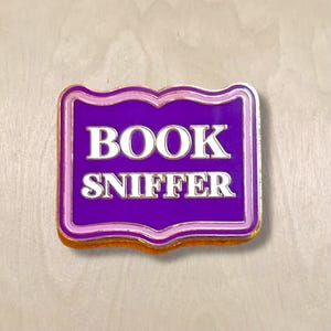 May include: A rectangular enamel pin with a purple background and the words "BOOK SNIFFER" in white capital letters. The pin has a scalloped edge with a pink and gold border.