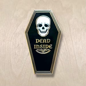 May include: A black coffin-shaped pin with a gold border and a white skull at the top. The words "DEAD INSIDE" are in gold lettering below the skull, with a decorative flourish beneath.