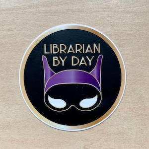 May include: A round sticker with a gold border and a black background. The sticker features a purple and white mask with the text "LIBRARIAN BY DAY" in gold letters.