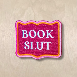 May include: A rectangular patch with the words "BOOK SLUT" embroidered in white and light blue on a magenta background. The patch has a decorative border with orange and pink accents. The patch is likely for a book lover.