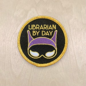 May include: A round, black embroidered patch with gold stitching. The patch features a purple and black superhero mask with the text "Librarian by Day" in gold.