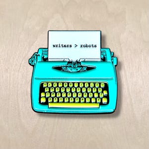 May include: A teal typewriter pin with a white paper insert. The paper reads "writers > robots" in black text. The typewriter has yellow keys and black lettering.