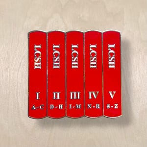 May include: A pin featuring five red book spines with the text "LCSH" in silver. Each book is labeled with a Roman numeral and a range of letters. The pin has a silver border.