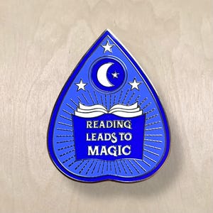 May include: A blue, teardrop-shaped pin with a gold border. The pin features a white crescent moon and stars at the top, with the words "READING LEADS TO MAGIC" in white text over an open book. The design has a radiating pattern.