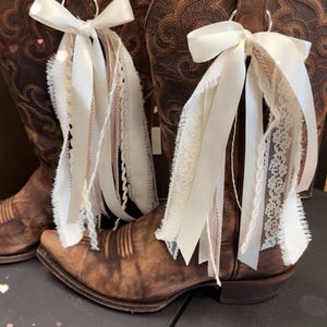 May include: A pair of brown leather cowboy boots with decorative stitching. Each boot is adorned with a white ribbon bow and cascading ribbons of lace and fabric in shades of white and pink. The boots are displayed on a dark surface.