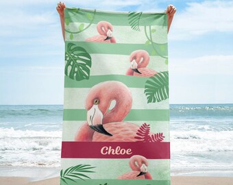 Op de afbeelding: Een groene en witte gestreepte strandhanddoek met een roze flamingo-ontwerp en groene tropische bladeren. De handdoek heeft de naam "Chloe" gedrukt op een rode streep.