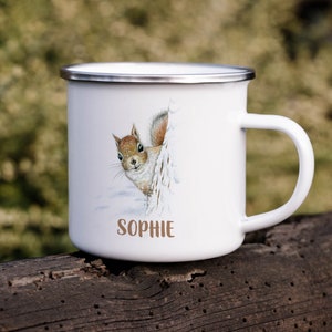 Puede incluir: Taza de esmalte blanco con una ardilla marrón asomándose desde detrás de un árbol nevado. La taza tiene un borde y un asa plateados y el nombre "Sophie" impreso en la parte inferior.