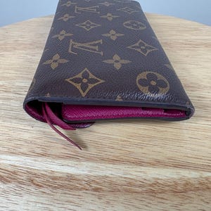 May include: Brown and tan wallet with a repeating pattern of floral and logo designs. The interior is a deep pink color. The wallet is made of a durable material and has a long, rectangular shape.