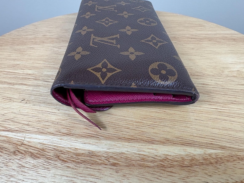 May include: Brown and tan wallet with a repeating pattern of floral and logo designs. The interior is a deep pink color. The wallet is made of a durable material and has a long, rectangular shape.