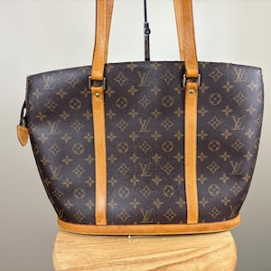 May include: Brown tote bag with the iconic LV monogram pattern. The bag features tan leather handles and trim, with a structured design. The bag has a rounded shape and a zipper closure. The bag is sitting on a wooden surface.