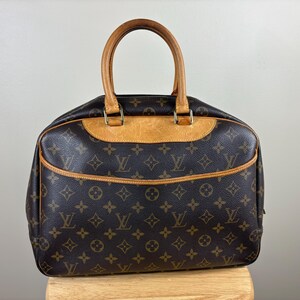 May include: Brown handbag with a repeating monogram pattern. The bag has a light brown leather trim and handles. The handles are curved and attached to the bag with gold-colored hardware. The bag is sitting on a wooden surface.