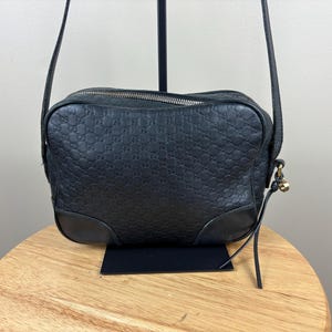 Puede incluir: Un bolso bandolera de cuero negro con un patrón de logotipo GG repetido en relieve en el exterior. El bolso rectangular tiene un cierre de cremallera superior, ribetes de cuero liso en la base y una correa de hombro fina con un pequeño dije de metal dorado.
