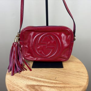 May include: A red leather crossbody bag featuring a prominent embossed interlocking G logo on the front. The bag includes a matching red leather tassel attached to a gold-tone metal ring on the side and a thin shoulder strap.