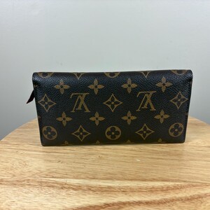 May include: A brown and tan wallet with a repeating pattern of floral and logo designs. The wallet is rectangular and appears to be made of leather. The wallet is sitting on a wooden surface.