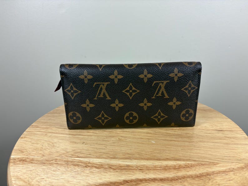 May include: A brown and tan wallet with a repeating pattern of floral and logo designs. The wallet is rectangular and appears to be made of leather. The wallet is sitting on a wooden surface.