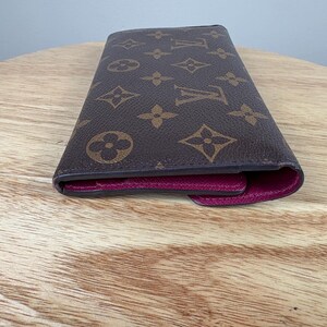 May include: Brown wallet with a repeating pattern of interlocking floral and geometric shapes. The interior is a deep magenta color. The wallet is closed and resting on a light brown wooden surface.