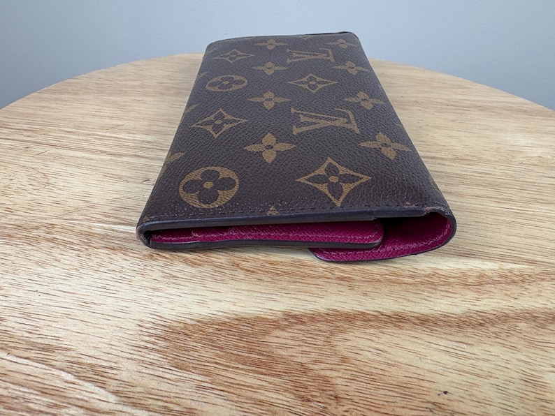 May include: Brown wallet with a repeating pattern of interlocking floral and geometric shapes. The interior is a deep magenta color. The wallet is closed and resting on a light brown wooden surface.