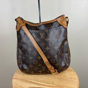 May include: Brown shoulder bag with a monogram pattern and a tan leather strap. The bag has a zipper closure and a front pocket. The bag is sitting on a wooden stool.