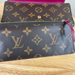 May include: Brown wallet with a gold zipper and a burgundy pull tab. The wallet features a repeating pattern of brown logos on a brown background. The interior of the wallet is a deep burgundy color.