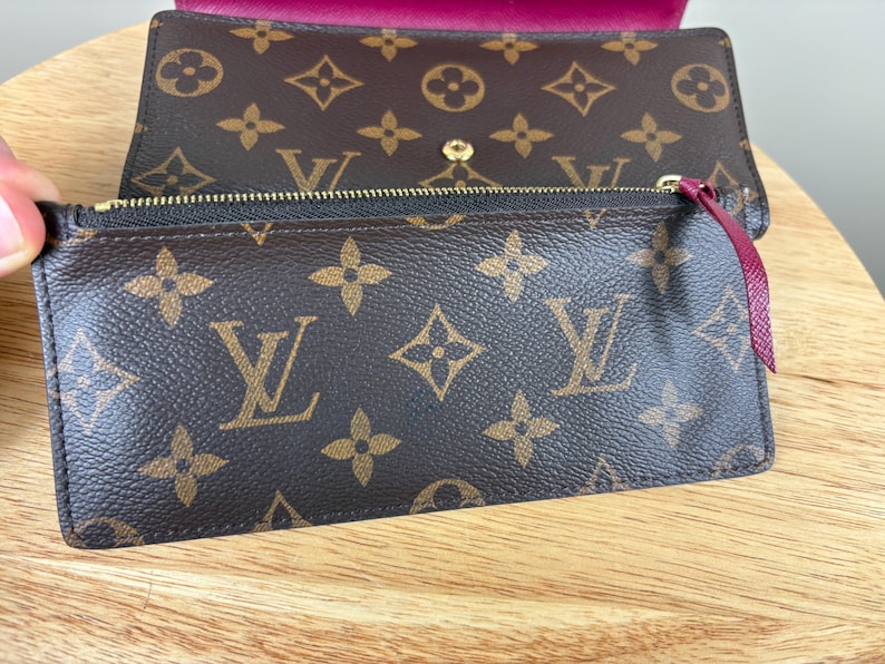 May include: Brown wallet with a gold zipper and a burgundy pull tab. The wallet features a repeating pattern of brown logos on a brown background. The interior of the wallet is a deep burgundy color.