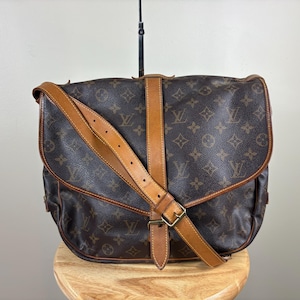 May include: Brown monogrammed messenger bag with tan leather trim and strap. The bag features a flap closure with a buckle and a shoulder strap. The bag is sitting on a wooden stool.