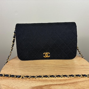 Puede incluir: Bolso de hombro negro acolchado con una correa de cadena dorada y un logotipo dorado. El bolso está hecho de un material suave y texturizado y se apoya sobre una superficie de madera. La correa de cadena está entrelazada con cuero negro.