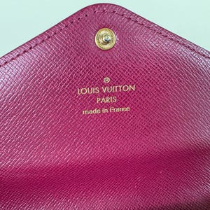 May include: Close-up of a magenta leather wallet with a gold snap closure. The interior features the embossed text "LOUIS VUITTON PARIS made in France" in gold lettering. The wallet has a textured surface.