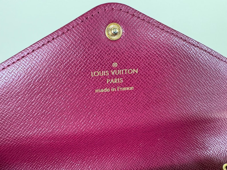 May include: Close-up of a magenta leather wallet with a gold snap closure. The interior features the embossed text "LOUIS VUITTON PARIS made in France" in gold lettering. The wallet has a textured surface.
