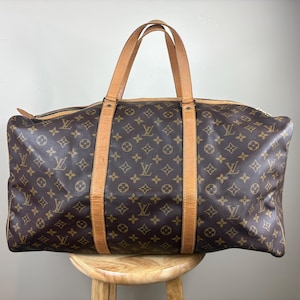 May include: A brown travel bag featuring the iconic Louis Vuitton monogram pattern in gold. The bag includes two tan leather handles that extend down the front and back, a top zipper closure, and a structured rectangular shape for storage.