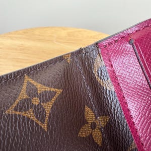 May include: Close-up of a brown leather wallet with a burgundy interior. The wallet features the iconic brown monogram pattern with the interlocking LV logo. The interior has card slots and a smooth, textured finish.