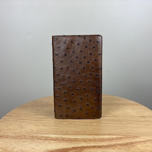 Puede incluir: Una cartera de cuero marrón con un patrón texturizado y punteado. La cartera es rectangular y se apoya sobre una superficie de madera. El cuero parece ser de un color marrón oscuro intenso.