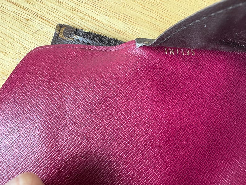 May include: Close-up of a magenta leather wallet interior. The wallet has a textured surface and the text "IN1195" is visible. A brown and tan patterned zipper is partially visible.
