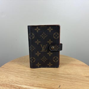 May include: A brown and dark brown leather planner cover featuring a repeating monogram pattern of flowers and the letters LV. The rectangular item includes a small side tab with a metallic snap button closure centered on the right edge.