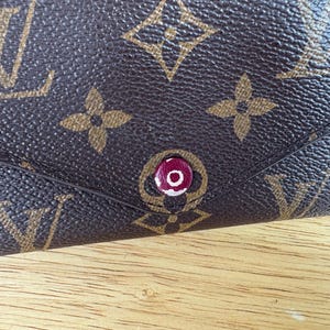 May include: Close-up of a brown leather wallet with a repeating pattern of interlocking logos and floral designs. The wallet features a red and white button closure. The background is a wooden surface.