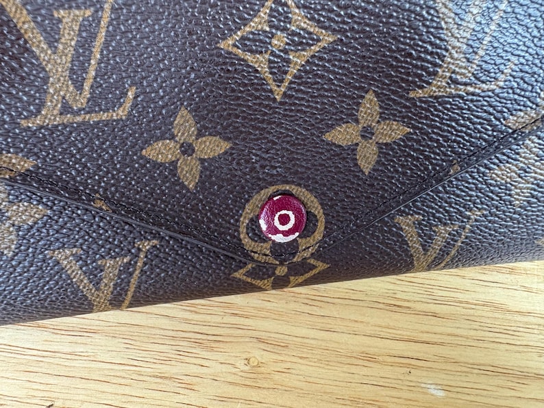 May include: Close-up of a brown leather wallet with a repeating pattern of interlocking logos and floral designs. The wallet features a red and white button closure. The background is a wooden surface.