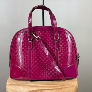 Puede incluir: Un bolso de mano en forma de cúpula de charol fucsia vibrante con un patrón de logotipo repetido en relieve. El bolso incluye dos asas superiores, una correa de hombro desmontable con herrajes plateados y una base lisa y sólida.
