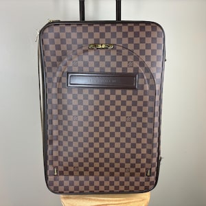 May include: Brown and tan checkered rolling suitcase with a retractable handle. The suitcase has a rectangular shape with rounded corners and a brown leather handle. The text "LOUIS VUITTON" is visible on the front.