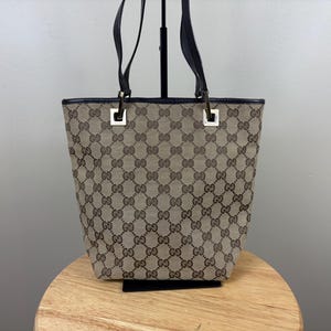 Puede incluir: Un bolso tote de lona en color beige y marrón con un patrón repetitivo del logotipo GG entrelazado. El bolso tiene asas de cuero negro sujetas con herrajes metálicos cuadrados de tono plateado en las esquinas superiores y borde de cuero.