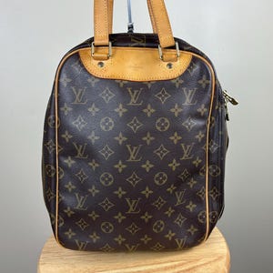 Puede incluir: Bolso de mano marrón con un patrón de monograma repetido. El bolso tiene asas y ribetes de cuero marrón, con cremallera y herrajes dorados. El bolso está sobre un taburete de madera.