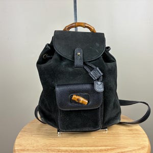 May include: Black suede backpack with a flap closure and a small front pocket. The backpack features a bamboo handle and a leather strap. The bag is sitting on a wooden surface.
