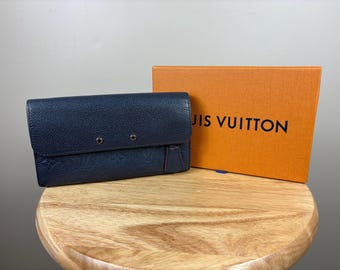 May include: A navy blue leather wallet with a flap closure and two silver-tone rivets. The wallet is next to an orange box with the words "LOUIS VUITTON" printed in black.