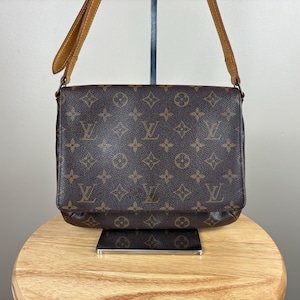 以下が含まれることがあります： モノグラム柄の茶色のショルダーバッグ。 フラップ開閉で、タンレザーのショルダーストラップが付いています。 バッグは木製の表面に置かれています。