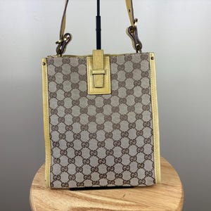 Puede incluir: Un bolso tote rectangular vertical con cuerpo de lona beige y un patrón repetitivo de logotipos GG marrones entrelazados. El bolso incluye ribetes de cuero amarillo claro en los laterales, una correa a juego y una lengüeta central de cuero.