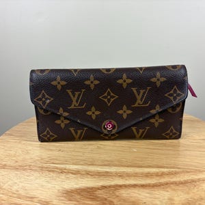 May include: Brown wallet with a flap closure, featuring a repeating pattern of brown floral and logo designs. The wallet has a pink accent and a snap closure. The wallet is sitting on a wooden surface.
