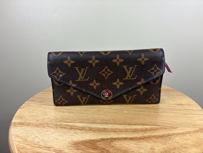 May include: Brown wallet with a flap closure, featuring a repeating pattern of brown floral and logo designs. The wallet has a pink accent and a snap closure. The wallet is sitting on a wooden surface.