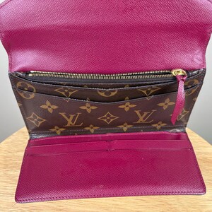 May include: Open, brown and pink wallet with a gold zipper and a pink pull tab. The wallet has a brown monogram pattern on the front and a pink interior with multiple card slots.