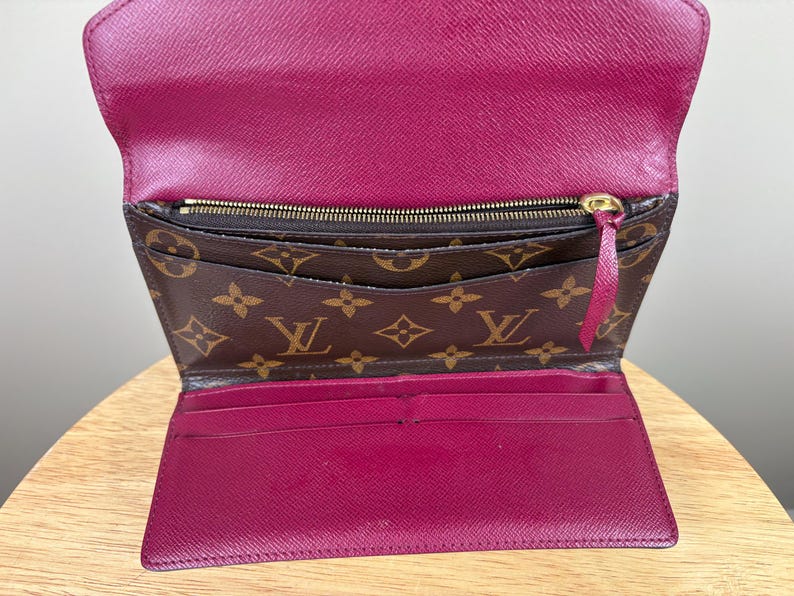 May include: Open, brown and pink wallet with a gold zipper and a pink pull tab. The wallet has a brown monogram pattern on the front and a pink interior with multiple card slots.