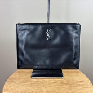 May include: Black leather clutch with a silver zipper and a silver YSL logo. The clutch is rectangular and appears to be made of smooth leather. The clutch is displayed on a wooden table.