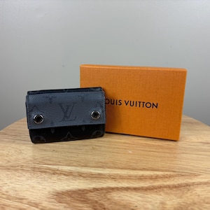 Może przedstawiać: Czarny i szary portfel z monogramem i srebrnymi elementami. Portfel znajduje się obok pomarańczowego pudełka z napisem "LOUIS VUITTON". Przedmioty znajdują się na drewnianej powierzchni.
