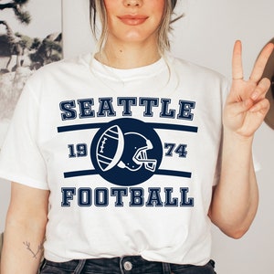 以下が含まれることがあります： 白地にネイビーブルーのグラフィックデザインが施されたTシャツ。"Seattle Football"の文字と、サークルの中にフットボールとヘルメットが描かれ、1974という数字が書かれている。
